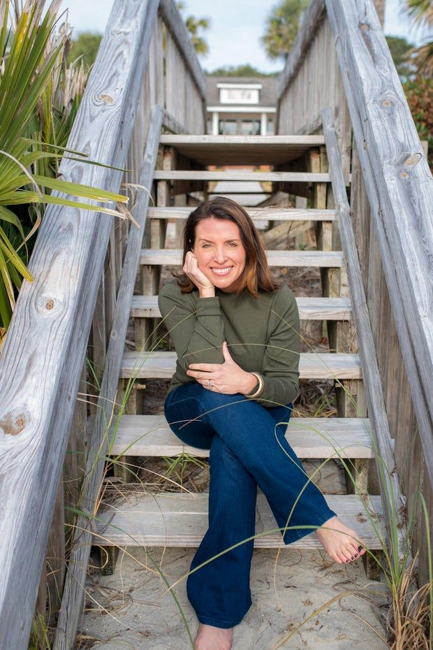 More women are drinking themselves sick. The Biden administration is concerned. 1 A smiling brown-haired woman sits on an outdoor staircase wearing a green sweater and jeans, her bare feet on the sand.